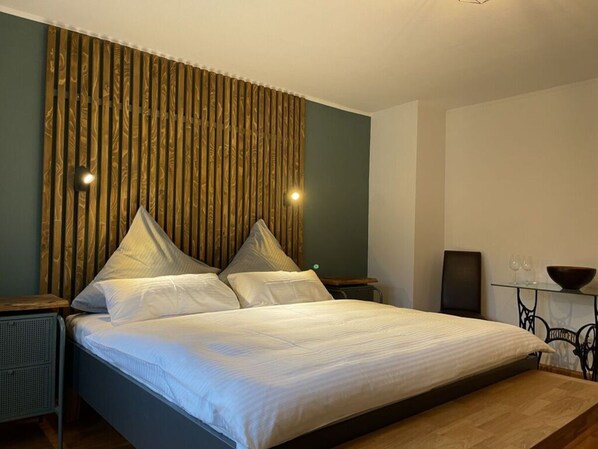 A neatly made bed with a headboard, a bedside table with a lamp, and a small table with glasses and a bowl.