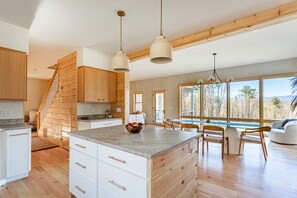 Een moderne keuken met houten kastjes, een groot eiland en een eetgedeelte met natuurlijk licht en uitzicht op de omgeving.