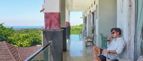 A balcony with a view of the sea, a person sitting on a chair, and a building with a tiled roof in the background.