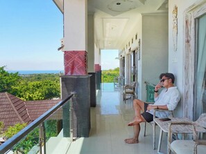 Ein Balkon mit Meerblick, eine Person sitzt auf einem Stuhl, und im Hintergrund ein Gebäude mit einem Ziegeldach.