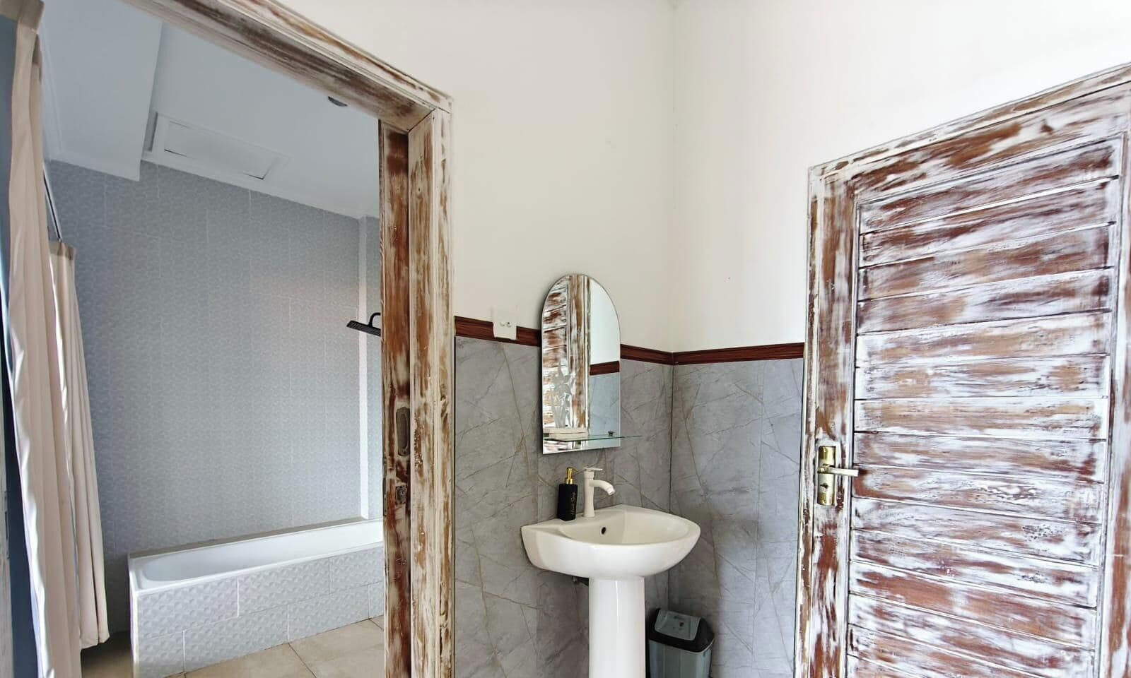 Un baño con lavamanos blanco, espejo y puerta con acabado de madera envejecida.