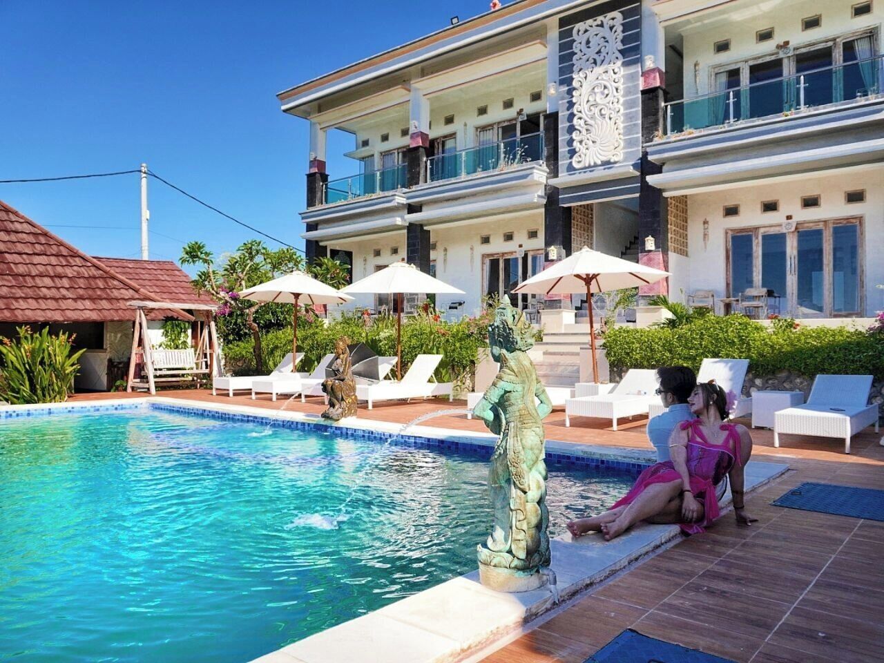 Una piscina con sillones de descanso y sombrillas, una estatua y un edificio con balcones.