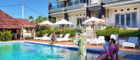 A swimming pool with lounge chairs and umbrellas, a statue, and a building with balconies.