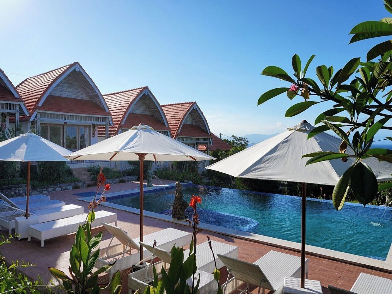 Zona de piscina con sillones y sombrillas, una pileta y edificios de estilo resort al fondo.