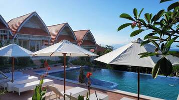 Zona de piscina con sillones y sombrillas, una pileta y edificios de estilo resort al fondo.