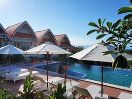 Zona de piscina con sillones y sombrillas, una pileta y edificios de estilo resort al fondo.