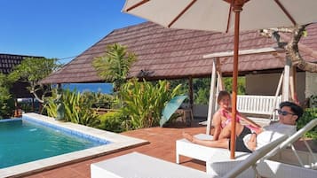 Un área junto a la piscina con sillones de descanso, un espacio de estar techado y vista al océano.