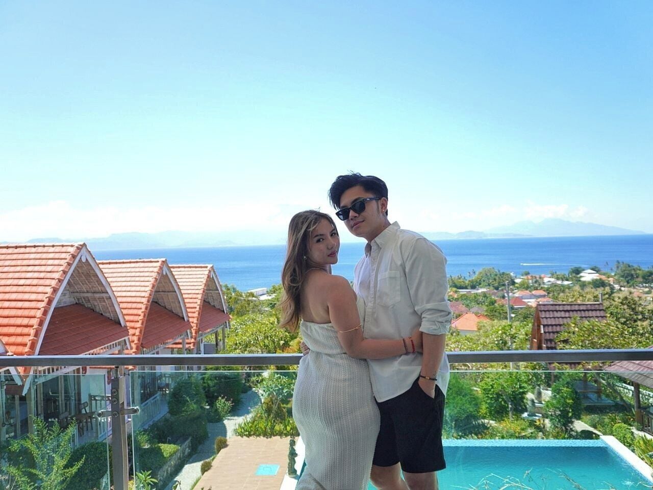 A couple standing by a pool with a view of the ocean and resort buildings.