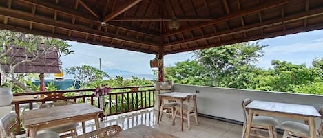 A covered outdoor seating area with wooden tables and chairs, a woven ceiling, and a view of greenery and a body of water.