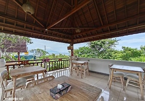Un'area salotto all'aperto coperta, con tavoli e sedie in legno, un soffitto intrecciato e vista su vegetazione e specchio d'acqua.