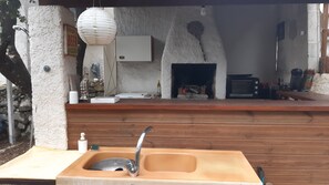 A rustic outdoor kitchen with a sink, a hanging light, and a stone fireplace.