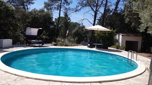 Une piscine dotée d’une terrasse carrelée, entourée d’arbres et bordée d’un petit bâtiment.