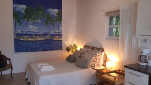 Ein Schlafzimmer mit Bett, Kissen, einem Nachttisch, einer Lampe, einer Kaffeemaschine, einem Fenster und einem Bild mit Strandmotiv.
