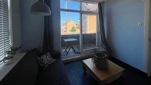 A modern living room with a sofa, a wooden coffee table, and a large window offering a view of buildings outside. - Unique House Within Walking Distance of the Beach (Katwijk Aan Zee)