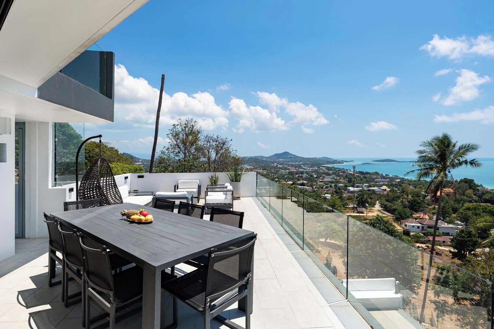 A modern outdoor dining area with a table and chairs, overlooking a coastal landscape with houses and a view of the sea.