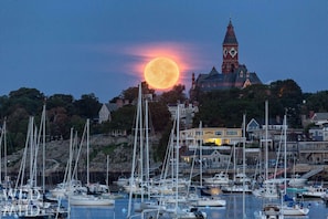 A harbor with multiple sailboats docked, a prominent clock tower, and a backdrop of a large moon. - Sailors Aweigh: Marblehead 3BR Retreat with Outdoor Space Near Harbor & Beaches (Marblehead)