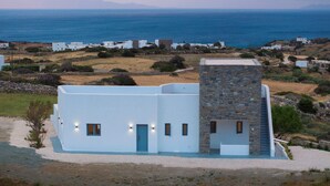 A modern white building with a stone tower, surrounded by a coastal landscape with houses in the background. - Morphe Villa (Aspro Chorio)
