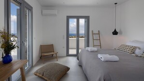 A modern bedroom with a large bed, a wooden chair, a small table with a vase, and a view through a glass door. - Morphe Villa (Aspro Chorio)