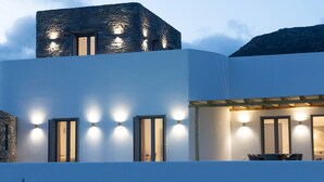 A modern building with a stone tower, multiple windows with lights, and a dining area with tables and chairs. - Morphe Villa (Aspro Chorio)