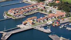 An aerial view of a coastal marina with multiple docked boats, a series of red-roofed buildings, and a grassy area with a few scattered trees. - 4 Person Holiday Park Home in Borre (Borre)