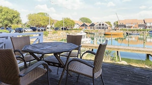 Une terrasse en bois, agrémentée de chaises et d’une table en osier, offre une vue sur une marina où l’on aperçoit des bateaux et des bâtiments en arrière-plan.