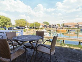 Una terraza de madera con sillas de mimbre y una mesa, con vistas a un puerto deportivo con barcos y edificios al fondo.