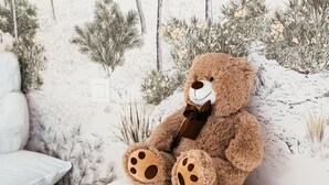 Un oso de peluche sentado en un sofá blanco frente a un mural con paisaje invernal.