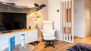 Una sala de estar moderna con un televisor de pantalla plana, un sillón blanco, una mesa de centro redonda y una original pieza de arte mural con forma de cola de ballena.