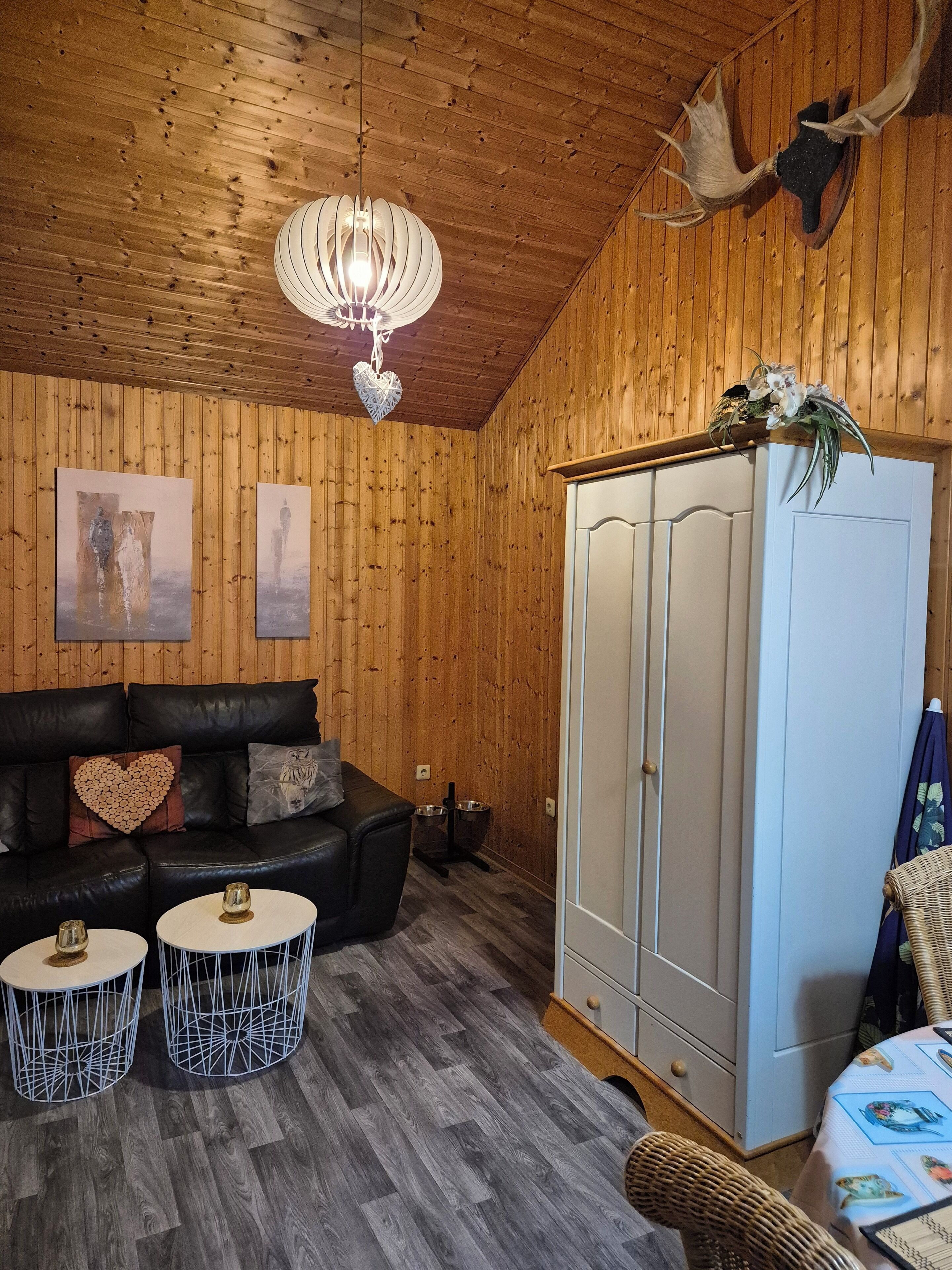 A wooden-paneled room with a black leather sofa, two white round tables, a white wardrobe, and a hanging deer antler.