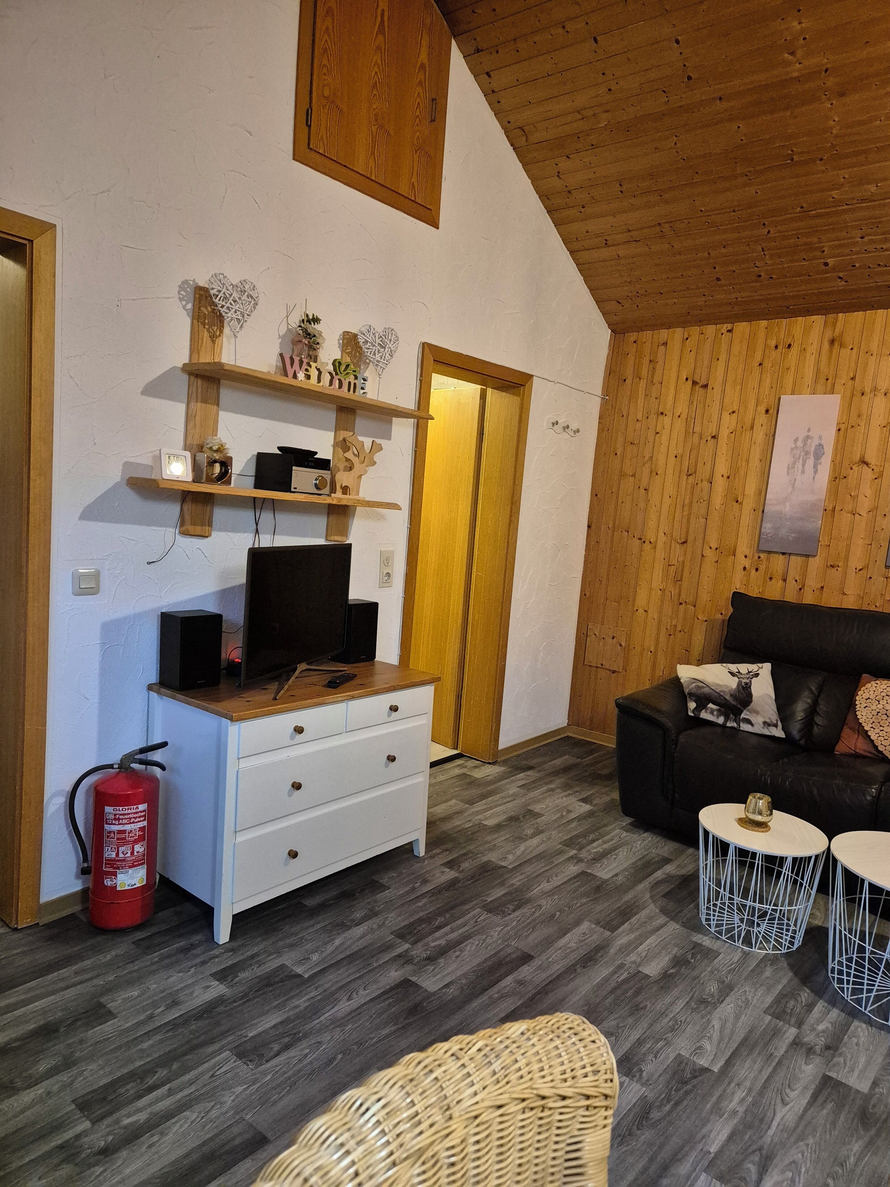 Een gezellige woonkamer met een houten plafond, een witte dressoir, een zwarte leren bank en een rieten stoel.
