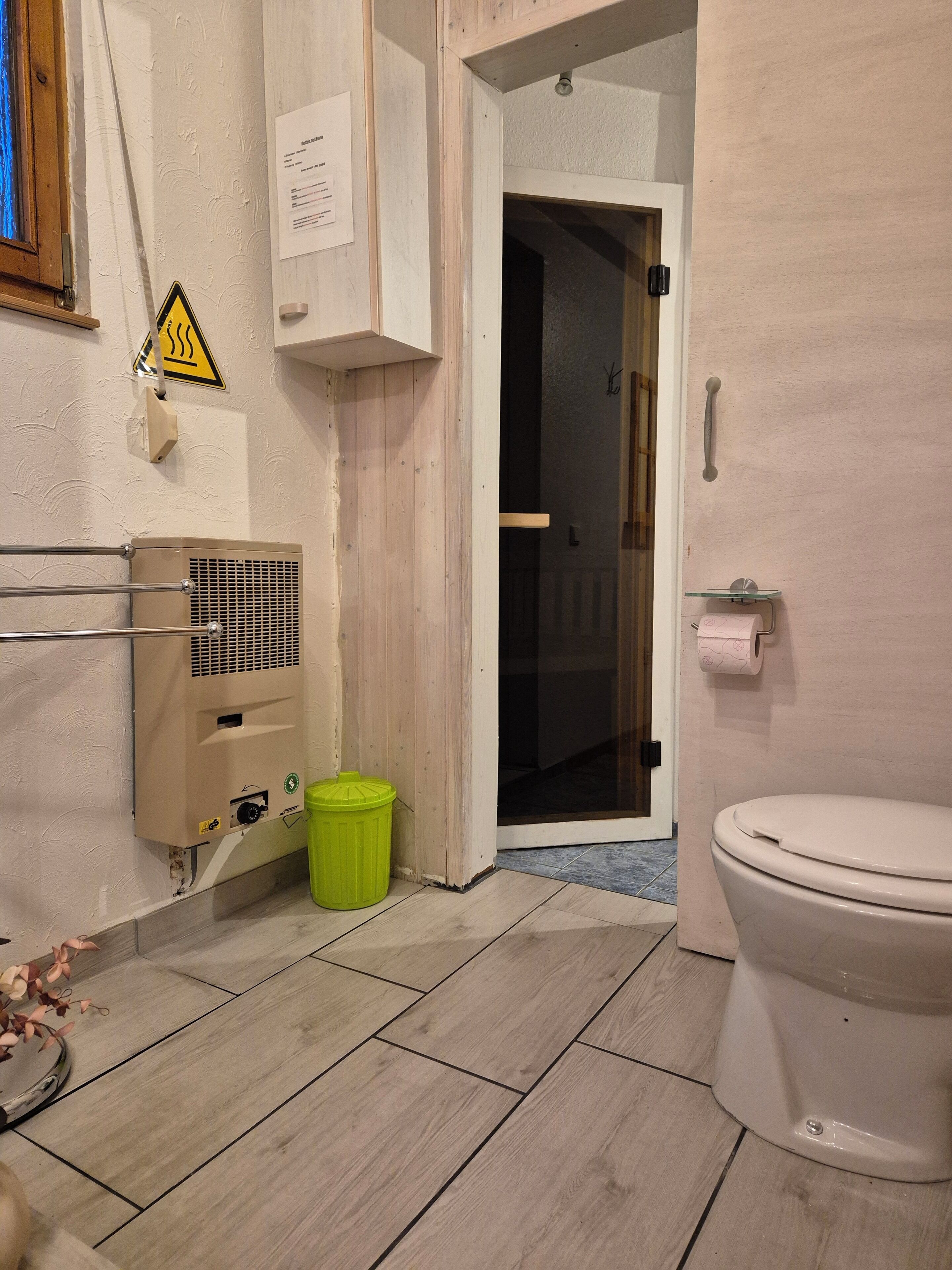 Un bagno con il water, un cestino verde, un dispenser per asciugamani di carta e un sistema di ventilazione a parete.