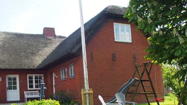 A red brick house with a thatched roof, a white door, and a number '185' on the wall. There is a white bench, a swing set, and a ladder leaning against the house. - Holiday Apartment Strandkrabbe (Ockholm)