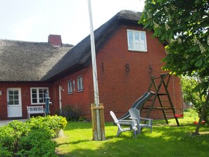 A red brick house with a thatched roof, a white door, and a number '185' on the wall. There is a white bench, a swing set, and a ladder leaning against the house. - Holiday Apartment Strandkrabbe (Ockholm)