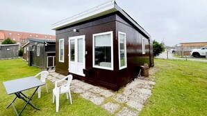 A small, dark-colored house with white-framed windows and a door, a patio with a table and chairs, and a grassy yard. - Lovely Holiday Home in Vlaanderen With Garden (De Haan)