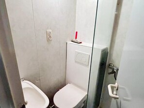 Casa de banho pequena e compacta, com duas sanitas, divisória de vidro e porta branca.