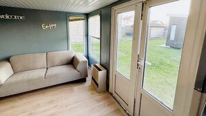 A room with a grey wall, a beige sofa, a wooden floor, and a large window with a view of a grassy yard. - Lovely Holiday Home in Vlaanderen With Garden (De Haan)