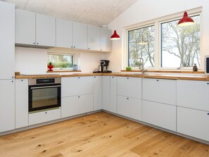 Une cuisine moderne avec des armoires blanches, un comptoir en bois, un four encastré, une fenêtre donnant sur les arbres et un luminaire suspendu rouge.