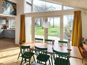 A dining area with a large table and chairs, a view of the outdoors through large windows, and a kitchen with cabinets and a sink. - 5 Star Holiday Home in Sydals (Sydals)