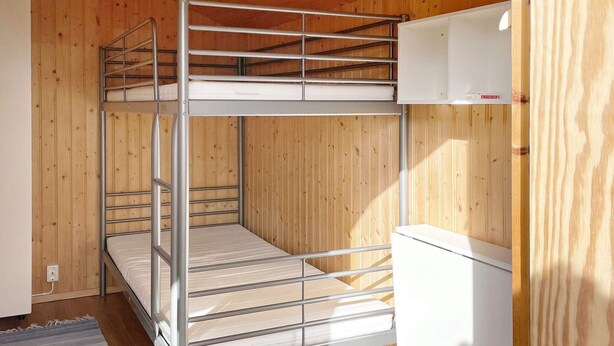 A bunk bed with a metal frame and white bedding, a wooden wall, and a white cabinet with a red label.