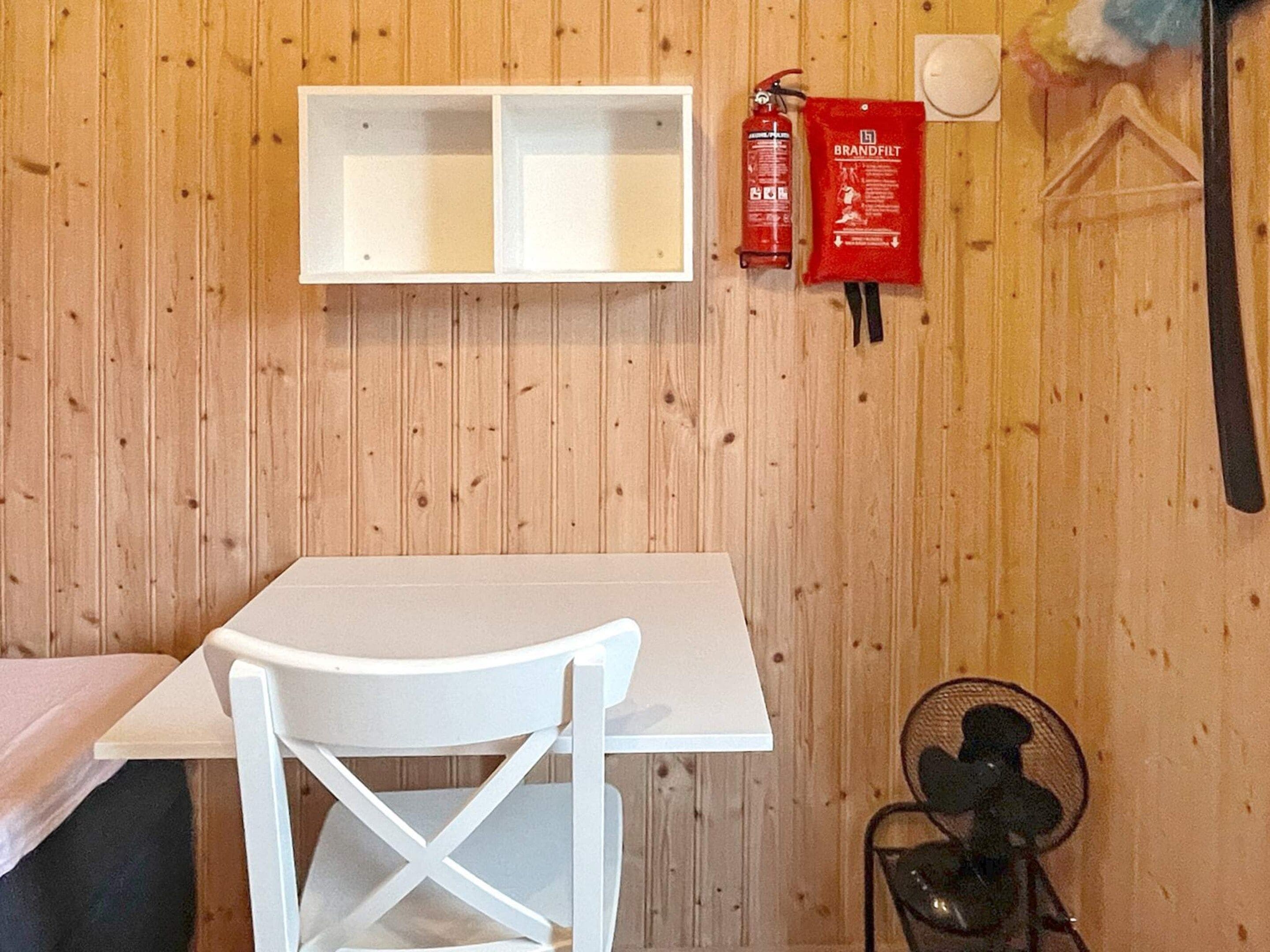 A room with a white table, a white chair, a fire extinguisher, a fan, and a shelf unit.