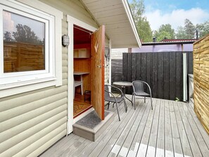 Une terrasse en bois avec une petite table et des chaises, un espace extérieur couvert et une porte coulissante donnant sur une pièce avec une fenêtre et une porte.