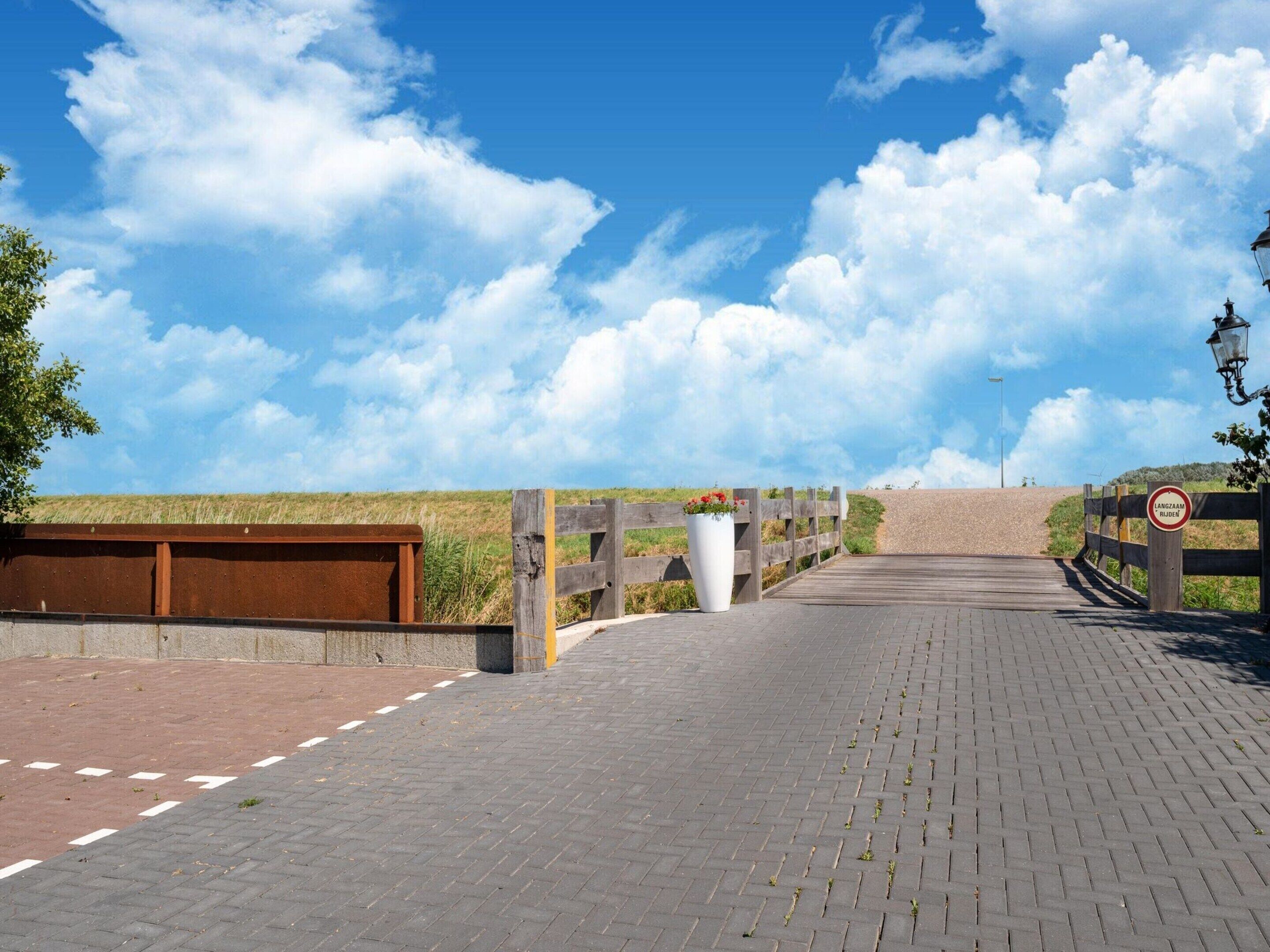 A paved pathway leading to a wooden gate with a sign that reads "Langzaam rijden" (Drive slowly).