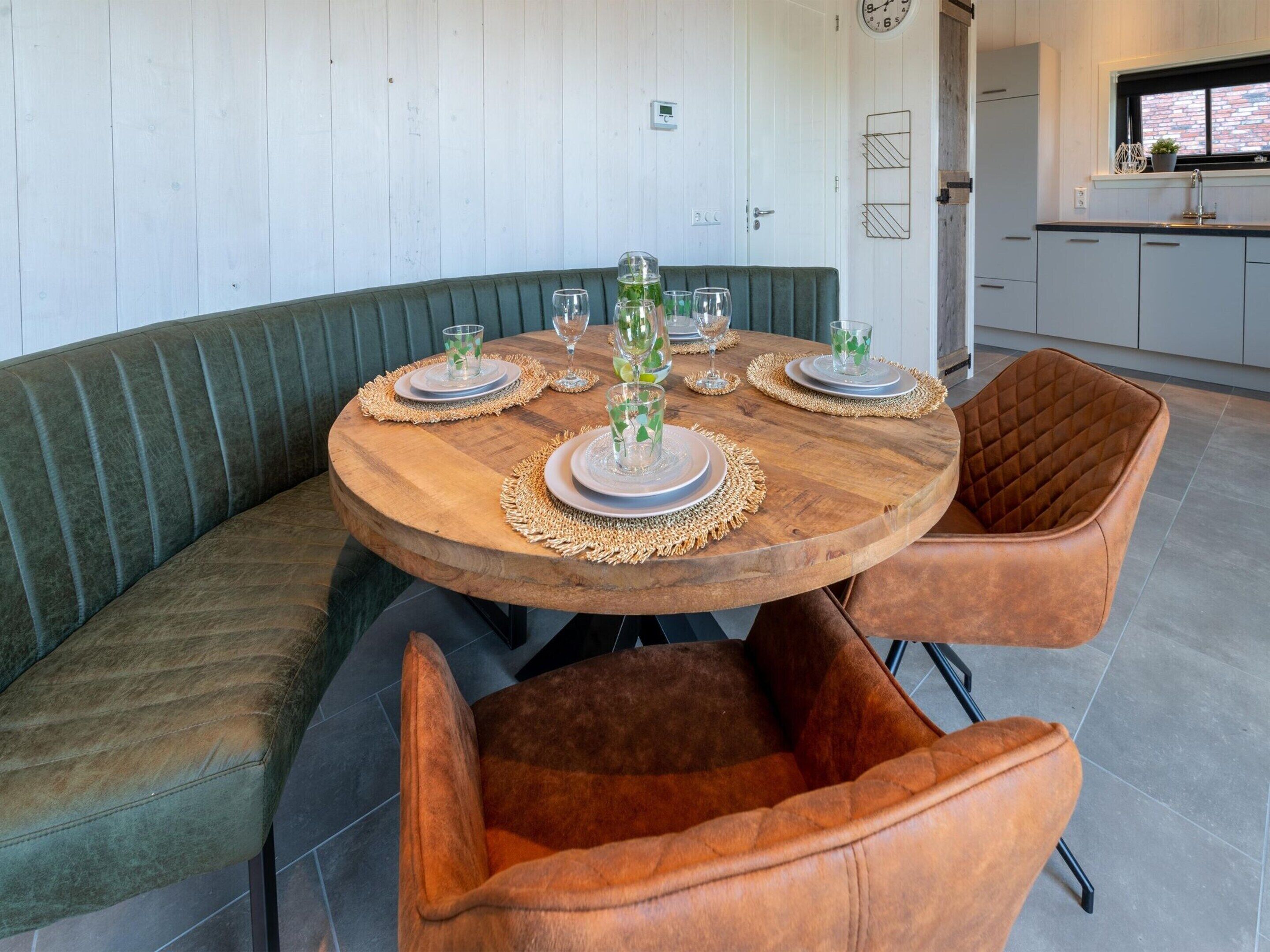 Een ronde houten tafel is gedekt met borden, glazen en een middelpunt. Er staan twee beklede stoelen met een gewatteerd patroon bij. De kamer heeft een houten bank langs de muur en een keuken met kastjes en een raam.