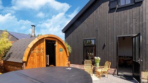 A rustic wooden building with a large arched entrance, a covered outdoor seating area with wicker chairs, and a car parked in front.