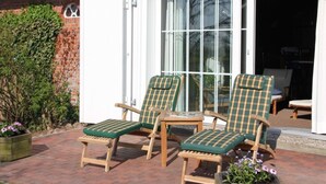 A patio with two wooden lounge chairs with green cushions, a brick floor, and a white building with a glass door. - Apartment in Ganzlin (Klanxbuell)