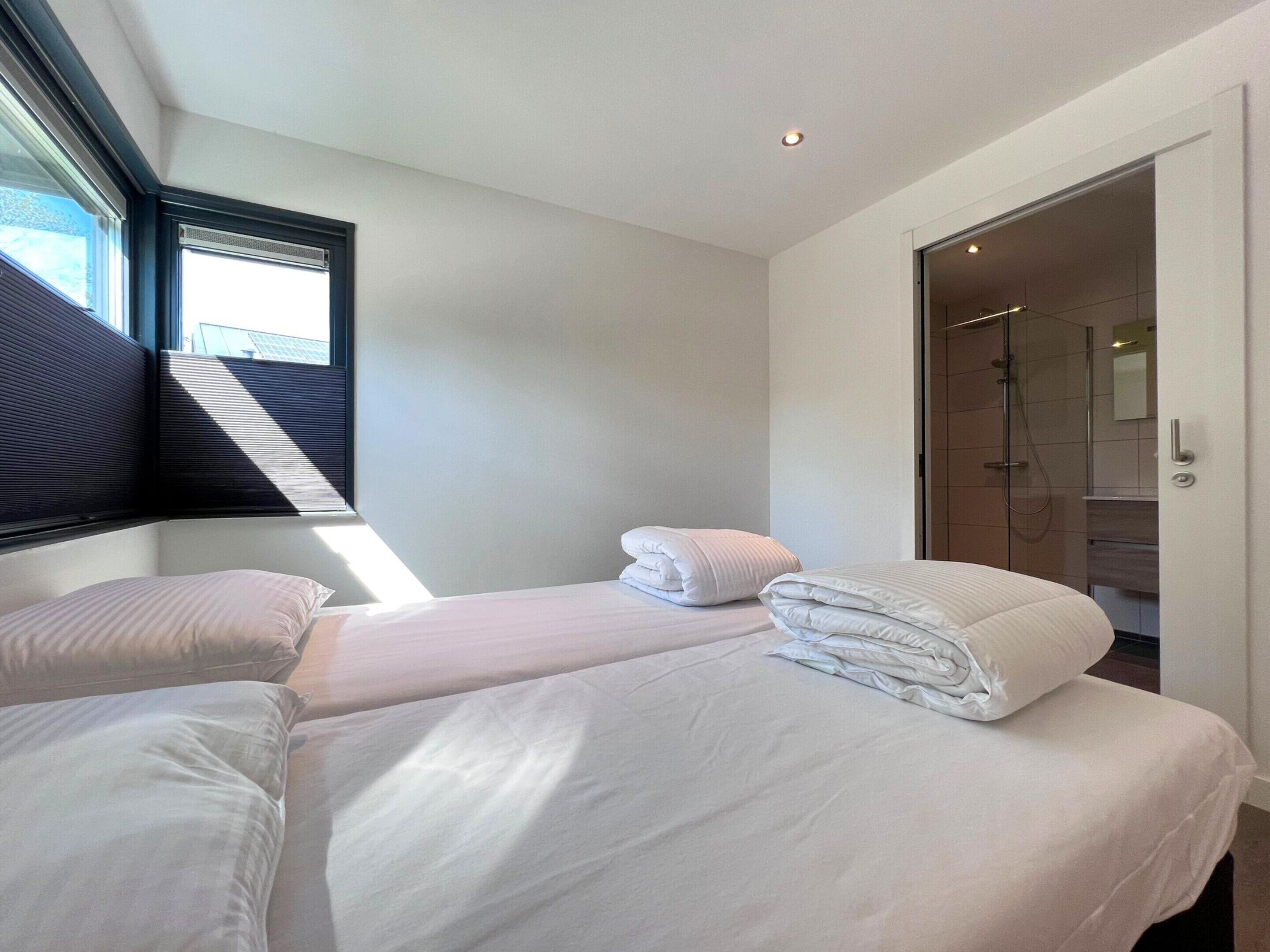 A modern hotel room with two beds, a large window, and a walk-in shower.