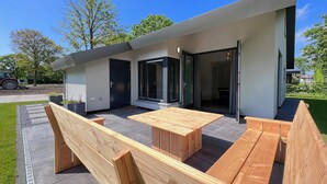 A modern outdoor patio with wooden benches and a table, a sliding glass door, and a small garden area. - Eco-friendly Villa With Terrace and Garden (Dirkshorn)