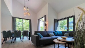 A modern living room with a dark grey sofa, a round coffee table, and a dining area with chairs and a table. Large windows provide natural light and views of greenery outside. - Eco-friendly Villa With Terrace and Garden (Dirkshorn)