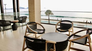 A balcony with wicker chairs and a round table, overlooking a beach with people and a palm tree.