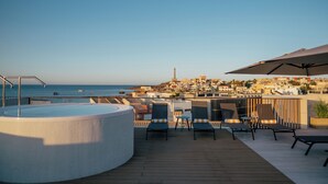 A rooftop terrace with a hot tub, lounge chairs, and a view of the sea and cityscape.